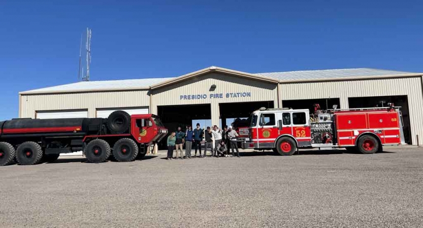 a group of students stand in front of a fire station and fire trucks while participating in a service project with outward bound